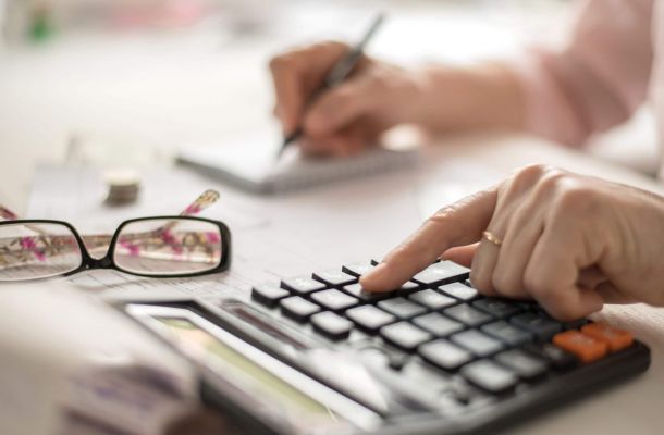 Hand einer Person tippt auf einem Taschenrechner, daneben liegen Unterlagen und eine Brille – symbolisch für die Berechnung von Pflegekosten und finanzielle Planung bei Betreuung.