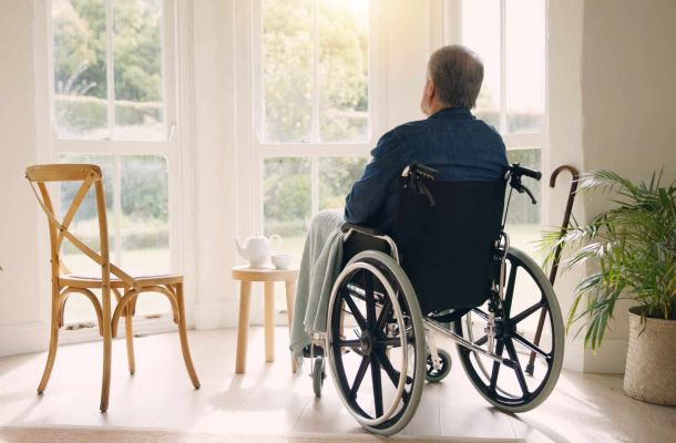 Älterer Mann im Rollstuhl sitzt in seinem Zuhause und blickt nachdenklich aus dem hellen Fenster.